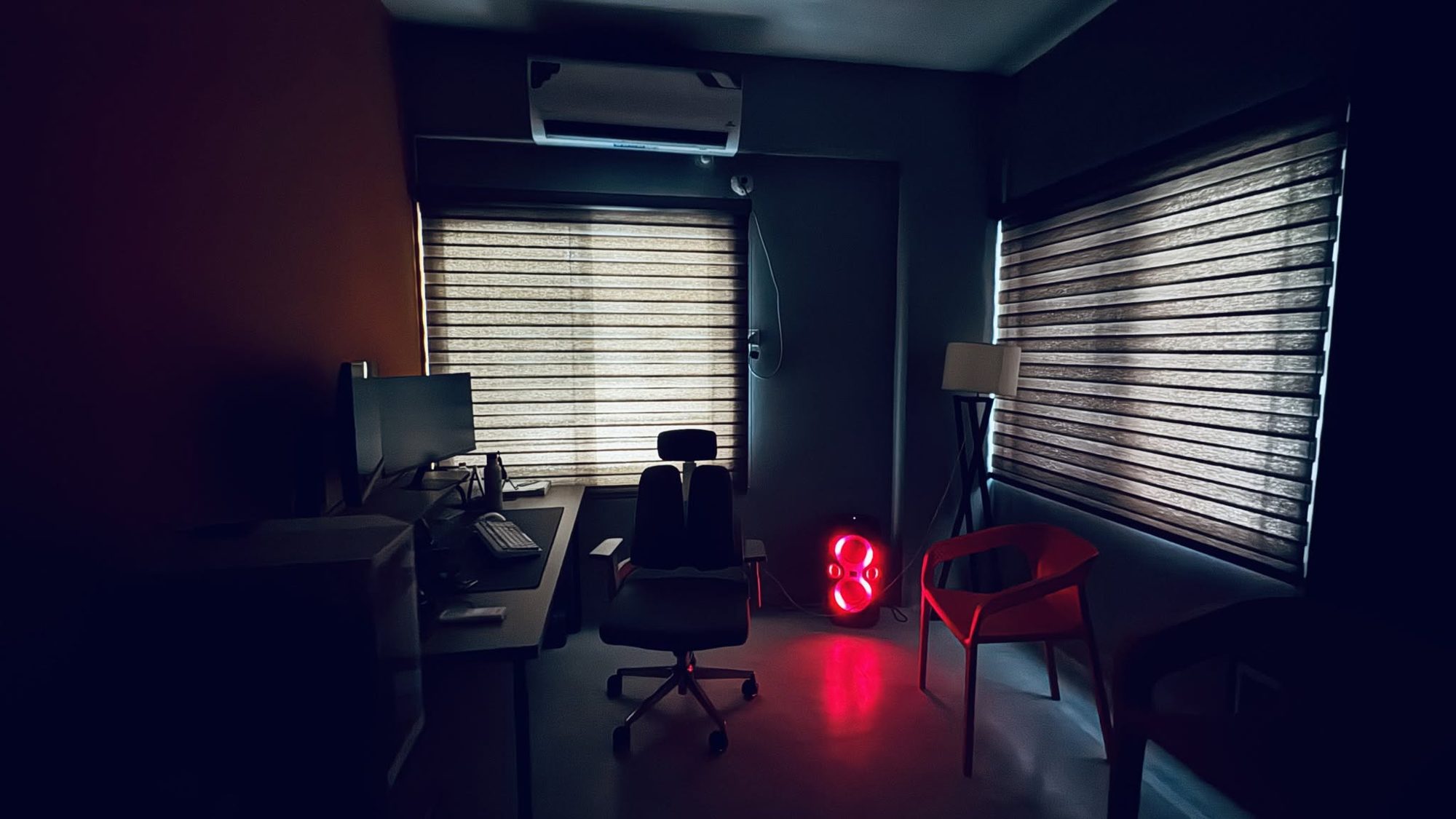 Monon's dimly lit studio room with a dual-monitor desk, office chair, glowing red speaker, floor lamp and red guest chair